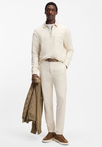 Beiger Pullover mit Viertel-Reißverschluss über einem weißen Hemd, cremefarbene Hose, braune Wildlederschuhe und eine khakifarbene gesteppte Jacke in der Hand. Glatte, neutrale Texturen.