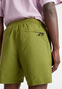 Pantalones cortos verdes con una cinturilla elástica, que cuentan con un pequeño bolsillo con cremallera en la parte trasera, fabricados con un tejido ligero y de textura suave.