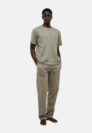 Homme debout, pieds nus, portant un t-shirt à manches courtes de couleur olive clair avec une poche poitrine, et un pantalon beige texturé, ample, avec cordon de serrage.