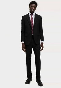 Schwarzer Anzug mit figurbetonter Silhouette, kombiniert mit einem weißen Hemd und einer rot gestreiften Krawatte. Die Schuhe sind aus schwarzem Leder.