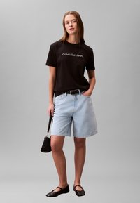 Czarna bawełniana koszulka z logo „Calvin Klein Jeans”, jasnoniebieskie szorty jeansowe, czarny pasek i płaskie czarne buty; trzyma małą czarną torebkę.