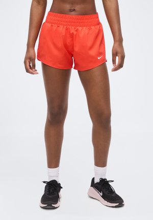 Mujer con pantalones cortos deportivos naranja brillante, calcetines blancos hasta el tobillo y zapatillas de correr negras y rosas, de pie sobre un fondo blanco.