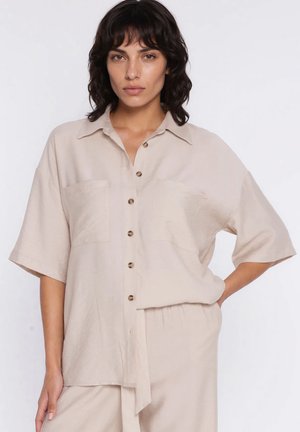 Kobieta o ciemnych włosach do ramion, ubrana w luźną, beżową koszulę zapinaną na guziki oraz pasujące spodnie, stojąca na białym tle.