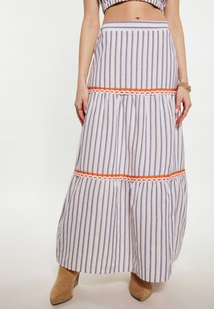 Vrouw draagt een gestreepte maxi rok met hoge taille en oranje kanten afwerking, gecombineerd met beige suède laarzen en een cropped top.