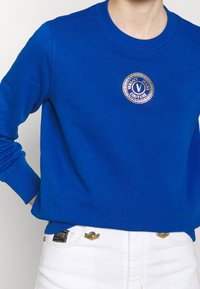 Blauwe katoenen sweatshirt met een ronde hals, geribbelde boorden en een ronde gouden logo patch met daarop "Versace Jeans Couture." Witte broek eronder.