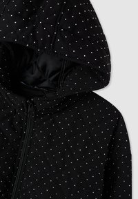 Giacca nera trapuntata con cappuccio, caratterizzata da piccoli punti argentati e chiusura con zip. Tessuto liscio con un leggero lucido e imbottitura.
