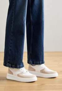 Baskets blanches avec des accents en suède beige, semelle plate et bout rond, associées à un jean large bleu foncé sur un sol en bois.