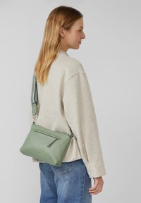 Borsa a tracolla in pelle verde con tasca frontale con zip e tracolla rimovibile, esposta su uno sfondo chiaro. Superficie testurizzata.