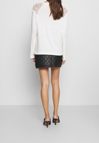 Donna con una maglietta bianca a maniche lunghe con spalline in pizzo, una mini gonna nera trapuntata e sandali neri con tacco alto, in piedi contro uno sfondo semplice.