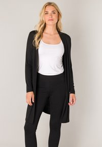 Zwarte lange cardigan van zacht stof, met een open voorkant en lange mouwen, gedragen over een wit tanktopje en zwarte legging.