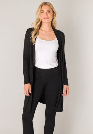 Zwarte lange cardigan van zacht stof, met een open voorkant en lange mouwen, gedragen over een wit tanktopje en zwarte legging.