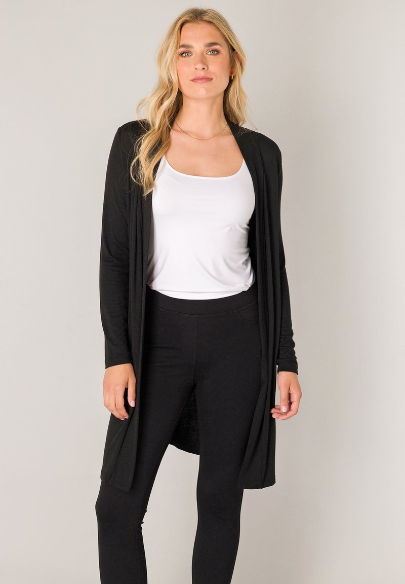 Zwarte lange cardigan van zacht stof, met een open voorkant en lange mouwen, gedragen over een wit tanktopje en zwarte legging.