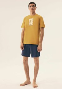 Złota bawełniana koszulka z grafiką "SUNRISE SURF CLUB". Połączona z granatowymi szortami. Miękka tekstura, okrągły dekolt, luźny krój.