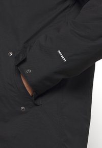 Veste imperméable noire avec tissu texturé, fermeture à pression, et logo "DRYVENT" sur la manche. Dispose d'une ouverture de poche latérale.