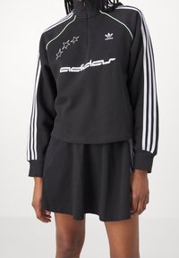 Svart zippered sweatshirt med vitt Adidas-logotyp och stjärndesign, med gröna accentlinjer och vita randiga ärmar, parat med en svart kjol.