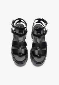 Black leather platform sandals featuring multiple crossed straps, adjustable ankle buckles, and a chunky, textured sole.