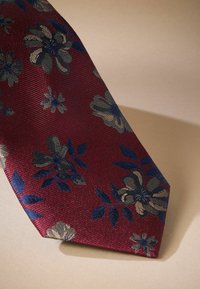 Cravate en soie bordeaux arborant un motif floral en bleu marine et or, avec une surface texturée et une pointe effilée, sur un fond neutre.