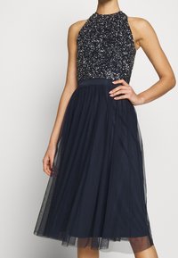 Laivaston mekko, jossa on kaula-aukko, paljeteista valmistettu yläosa ja tyllihelma. Tekstuuri yhdistää kimalluksen pehmeään, virtaavaan muotoiluun.