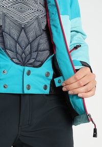 Bouton-pression à main sur une veste turquoise avec une fermeture éclair rouge, portant un pantalon noir et une chemise noire et blanche à motifs en dessous.