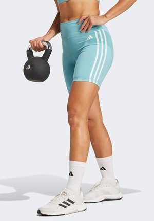 Kobieta w jasnoniebieskich szortach Adidas i białych tenisówkach trzyma czarny kettlebell z logo Adidas, stojąc na jasnej podłodze.