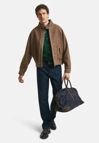 Bruine herringbone jas met een hoge kraag, groene shirt, blauwe denim broek en zwarte laarzen. Houdt een marineblauwe sporttas vast met bruine accenten.