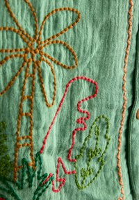 Tessuto verde con ricamo a mano arancione, rosso e verde raffigurante una palma e forme di foglie astratte.