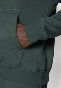 Felpa verde in pile con tasca a marsupio, polsini a coste e un accentuazione in pelle marrone. Presenta un piccolo logo Nike nero sul petto.