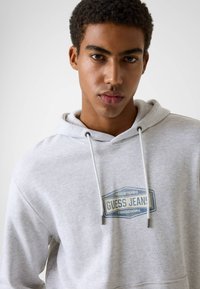 Szara bluza z kapturem, kieszenią typu kangur, sznurkiem do regulacji kaptura oraz niebieską, prostokątną naszywką z napisem "GUESS JEANS". Miękka, teksturowana tkanina.