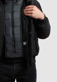 Schwarze Steppjacke mit Reißverschluss, Seitentaschen und gerippten Bündchen. Das Innenfutter ist gesteppte mit einem Logoschild.