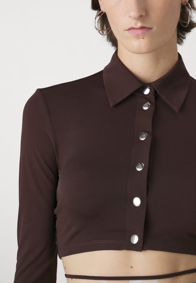Brown cropped shirt with a collar, long sleeves, front snap buttons, and smooth texture. Designed to fit snugly around the torso.