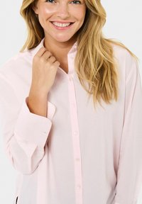 Femme souriante avec de longs cheveux blonds, portant une chemise boutonnée rose clair, tenant le col avec sa main droite.