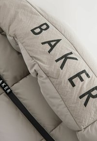 Kantet beige dyne med sort "BAKER" tekst, der har en glat overflade og kontrasterende sort stof langs kanten.