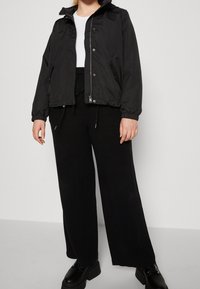 Chaqueta negra con cremallera frontal, capucha, puños elásticos y diseño ligeramente corto, combinada con pantalones negros holgados y zapatos negros gruesos.