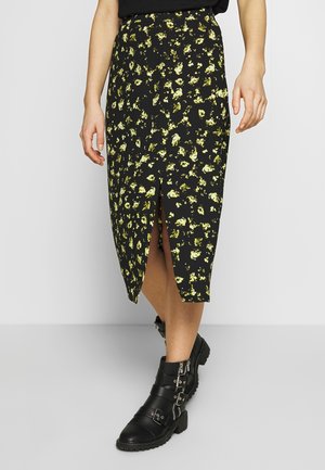 Sort midi-nederdel med gul blomsterprint og frontslids, båret med sorte ankelstøvler med spænder på en lys baggrund.