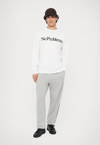 White long-sleeve shirt with "No Problemo" text in black, paired with light gray sweatpants featuring a side logo. Black sneakers complete the outfit.