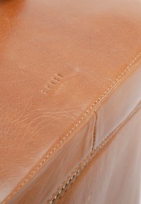 Ottomán de cuero marrón claro con una textura suave, costuras visibles y un branding sutil. Las esquinas redondeadas y un acabado pulido mejoran su diseño.