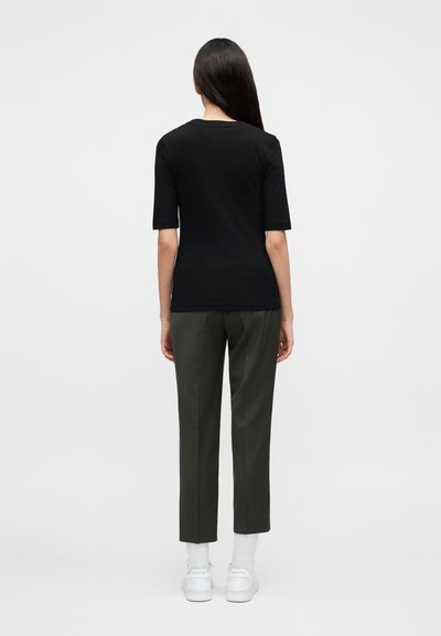 Γυναίκα με μακριά σκούρα μαλλιά, που φοράει μαύρο κοντομάνικο μπλουζάκι, σκούρο ψηλόμεσο παντελόνι μέχρι τον αστράγαλο, λευκές κάλτσες και λευκά αθλητικά παπούτσια, στέκεται μπροστά σε ένα απλό φόντο.