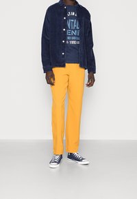 Mężczyzna w granatowej kurtce ze sztruksu, granatowej koszulce z grafiką, jaskrawożółtych spodniach oraz granatowych wysokich trampkach, stojący na szarej podłodze.