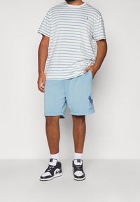 Pruhované tričko s krátkým rukávem v bílé a světle modré barvě, spárované se světle modrými šortkami s vyšitým logem, nošeno s černobílými teniskami.