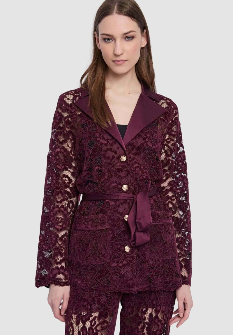 Blazer in pizzo color bordeaux con colletto in raso, maniche lunghe, bottoni dorati e cintura in vita. Presenta un intricato motivo floreale e tasche.