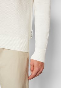Weißer Strukturpullover mit gerippten Bündchen, versehen mit einem gestickten Markenlabel an der Seite, kombiniert mit hellbeigen Hosen.