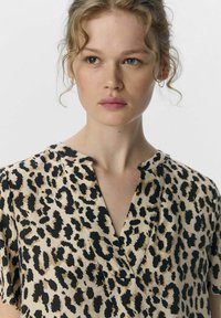 Blouse à imprimé léopard en beige et noir, avec un col en V, des manches courtes et une texture lisse. Détails de couture subtils visibles.