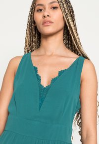 Vestido verde-água com um decote em V profundo, apresentando detalhes intrincados em renda. Design sem mangas e cintura ajustada que se transforma numa saia. Textura de tecido suave.