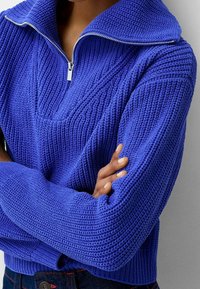 Pull-over bleu tricoté avec un col montant et une demi-fermeture éclair, présentant un motif texturé et des poignets côtelés. Le tissu semble épais et confortable.