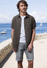 Braunes Kurzarmhemd aus strukturiertem Stoff mit Knopfverschluss, kombiniert mit hochgekrempelten grauen Denim-Shorts. Lässiges Sommer-Outfit am Meer.