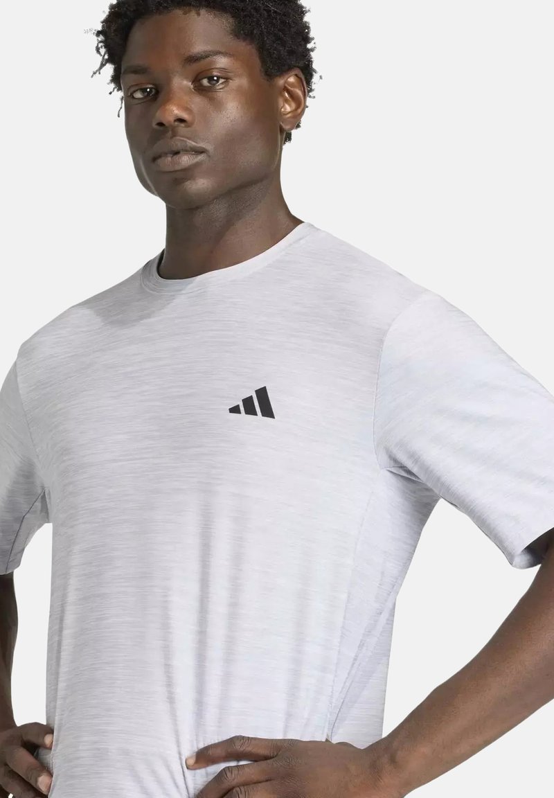 Homme aux cheveux courts et bouclés, portant un t-shirt blanc Adidas avec un logo sur la poitrine, mains sur les hanches, fond neutre.