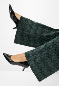 Tacchi in vernice nera con punta a punta, abbinati a pantaloni verdi larghi con una texture a motivi geometrici delicati.