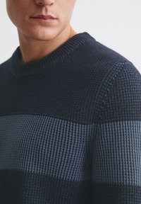 Marineblauer Pullover mit strukturiertem Strickmuster und horizontalen Streifen in hellerem Blau. Verfügt über einen gerippten Rundhalsausschnitt und enganliegende Ärmel.