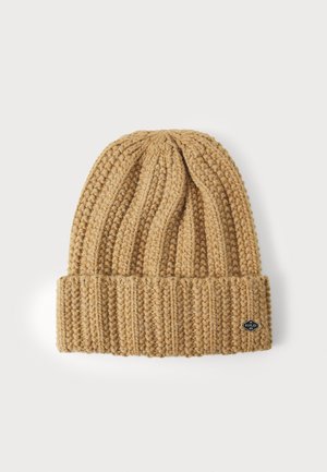 Brede gebreide beanie in tan, met ribbelstructuur, opvouwbare rand en een klein zwart logo op de zijkant.