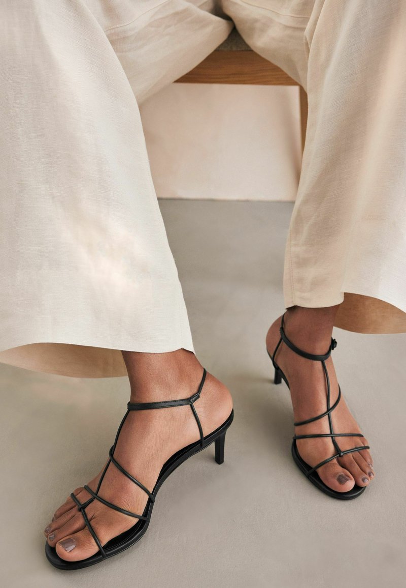 Svarta högklackade sandaler med remmar och minimalistisk design, med tunna remmar och fyrkantig tå, placerade mot beige tygbyxor.